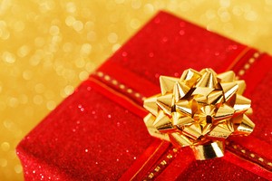 A zoomed-in photo depicting part of a holiday gift box. It is wrapped in glittery, red paper and topped with a golden bow.